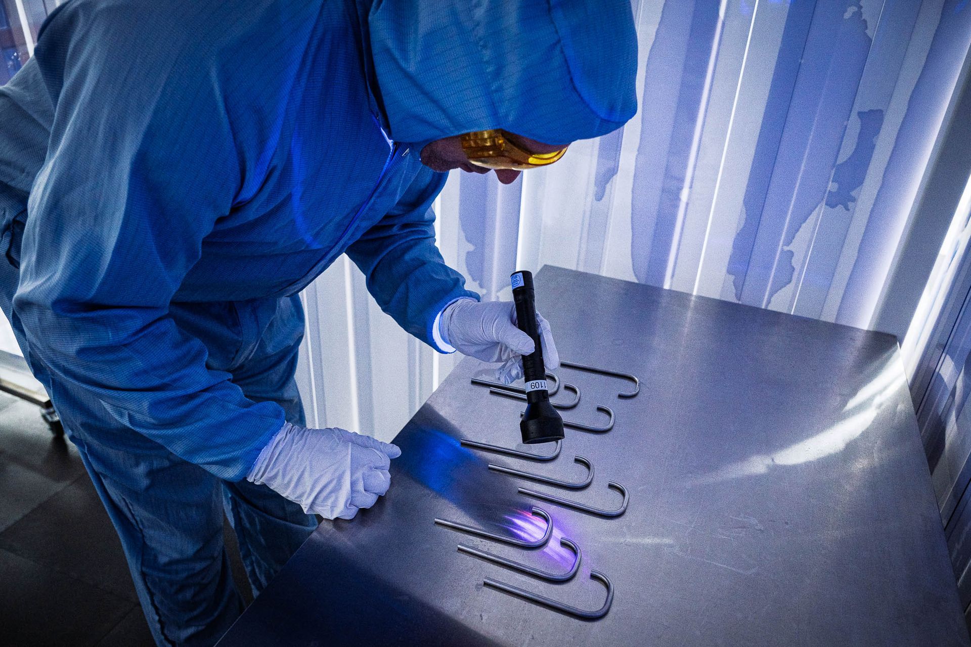 Een persoon in een blauw, schoon pak, handschoenen en een veiligheidsbril gebruikt UV-licht om metalen gereedschappen op een tafel te inspecteren.