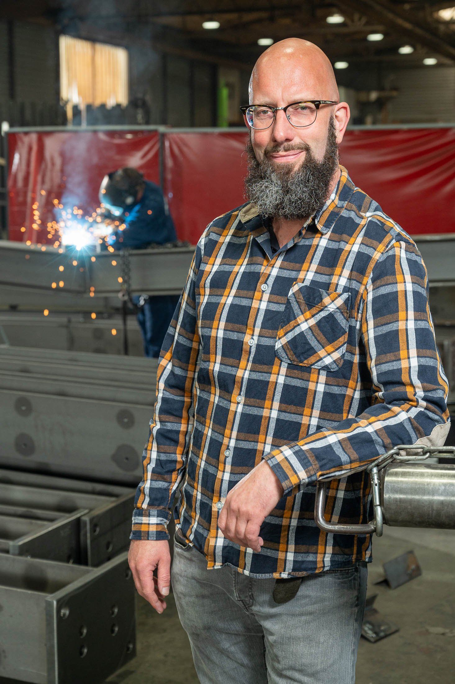 Een man met een baard en een bril staat voor een lasapparaat in een fabriek.