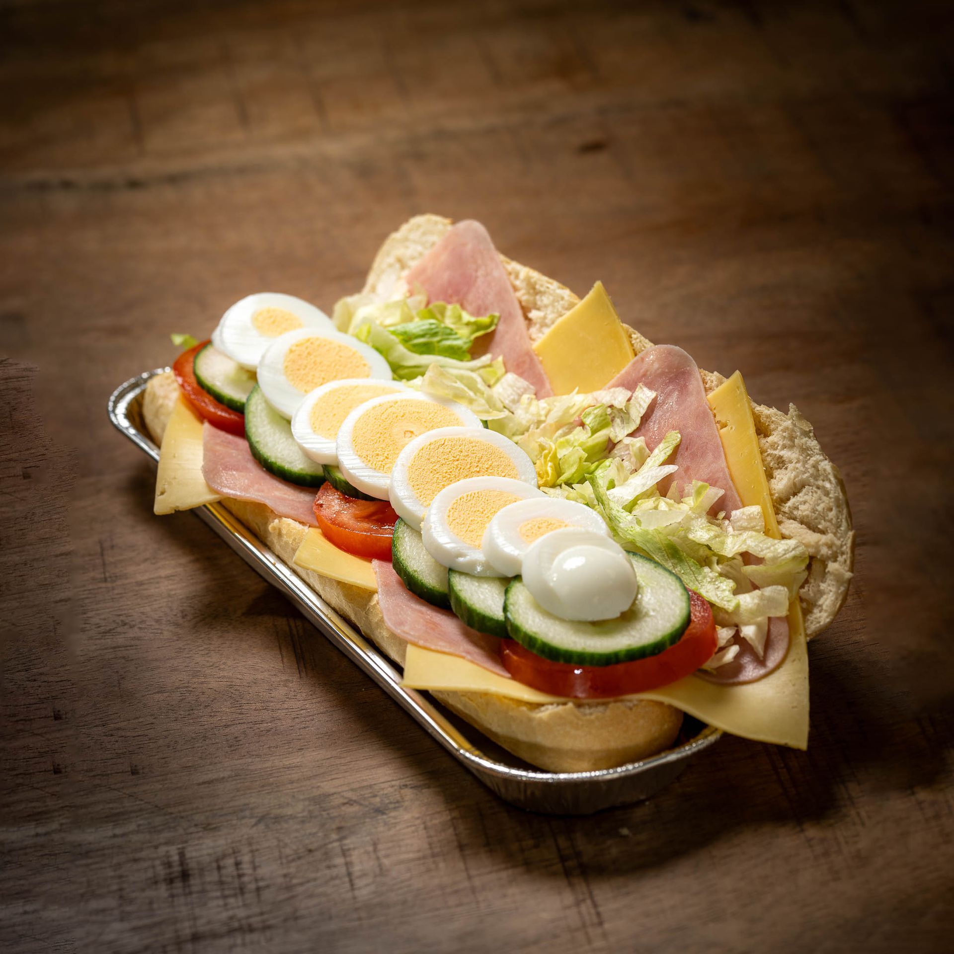 Een sandwich met ham, kaas, eieren, komkommers en tomaten op een houten tafel.