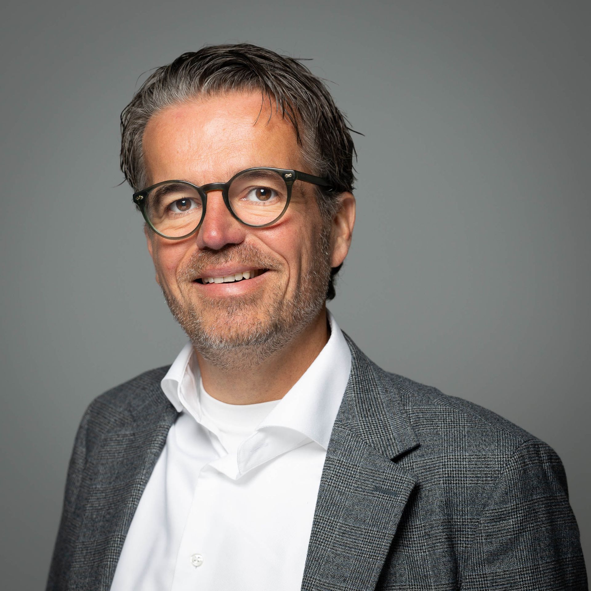 Een man met een bril, lachend, gekleed in een blazer en een wit overhemd, tegen een grijze achtergrond.