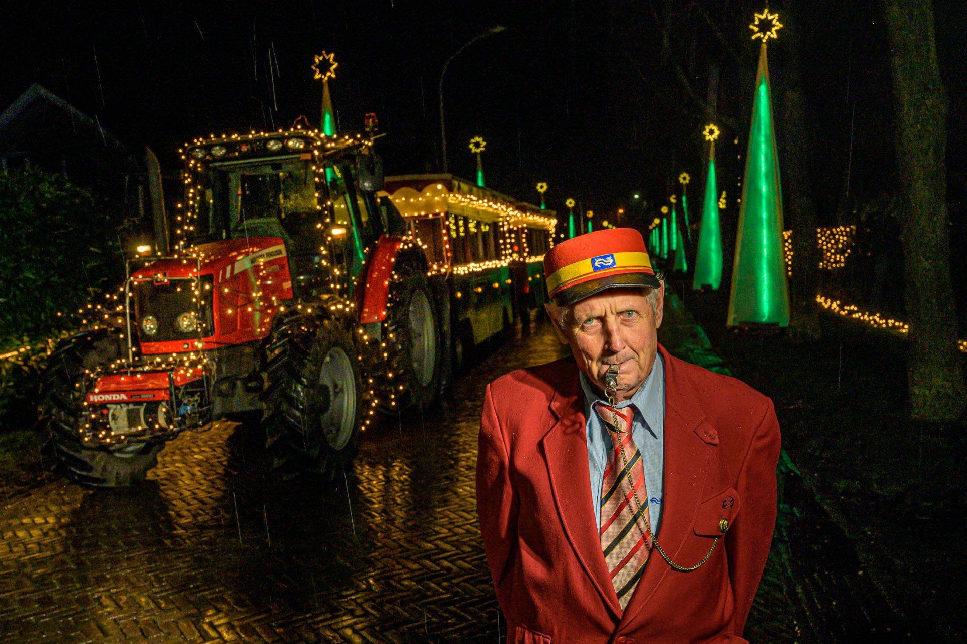 Een man in een rood pak en stropdas staat voor een tractor die versierd is met kerstverlichting.