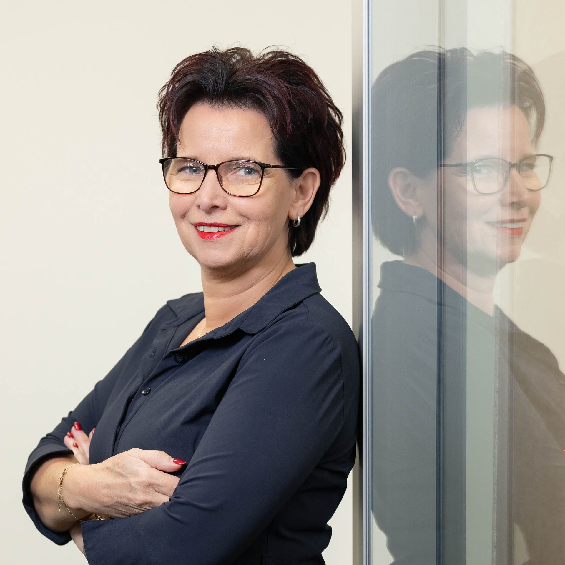 Een vrouw met een bril leunt tegen een muur met haar armen over elkaar.