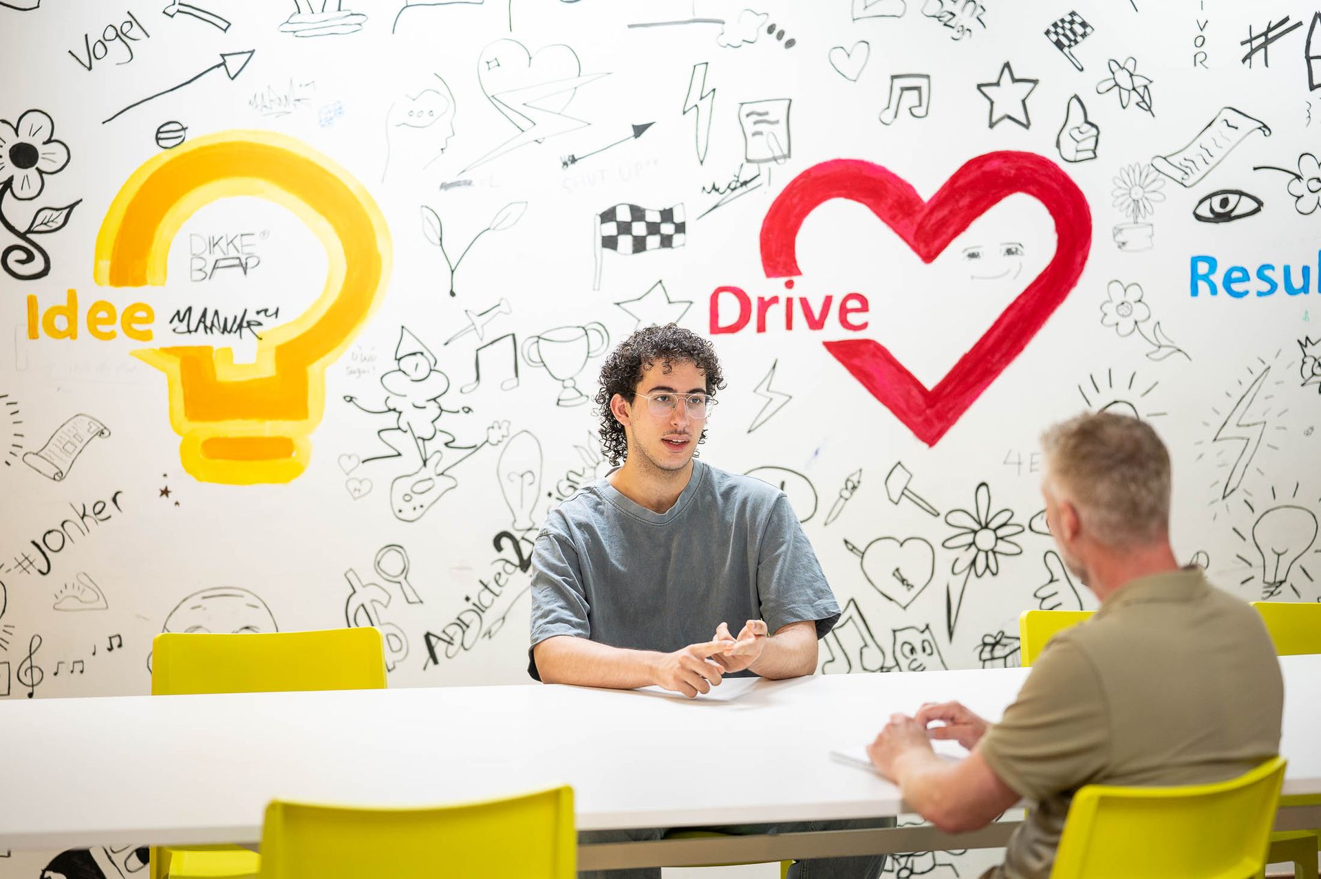 Twee mannen zitten aan een tafel en voeren een gesprek voor een muur vol tekeningen, waaronder een gloeilamp en een hart.