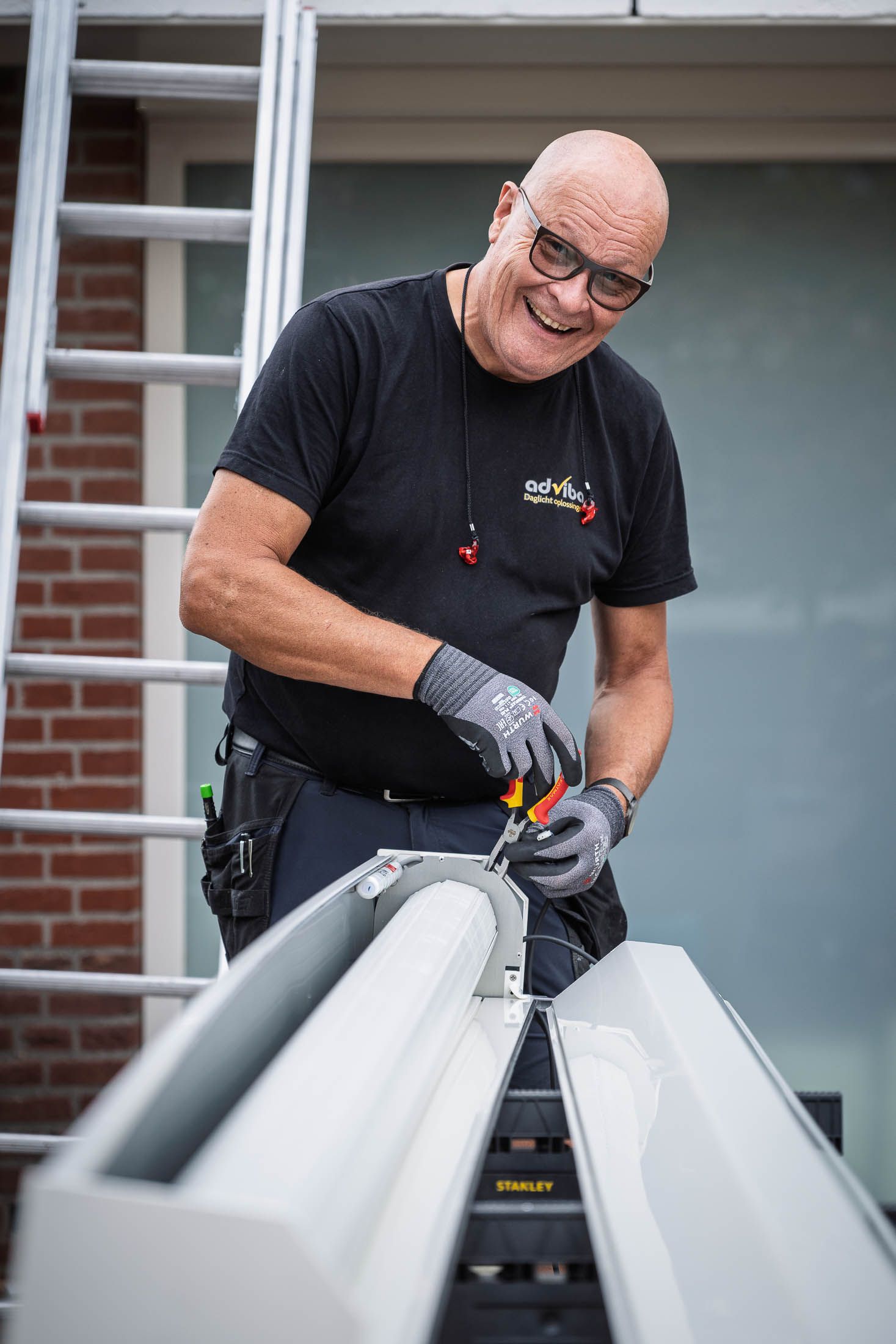 Man in zwart shirt met handschoenen gebruikt een schroevendraaier om een ​​witte dakgoot te monteren. Op de achtergrond is een ladder te zien.