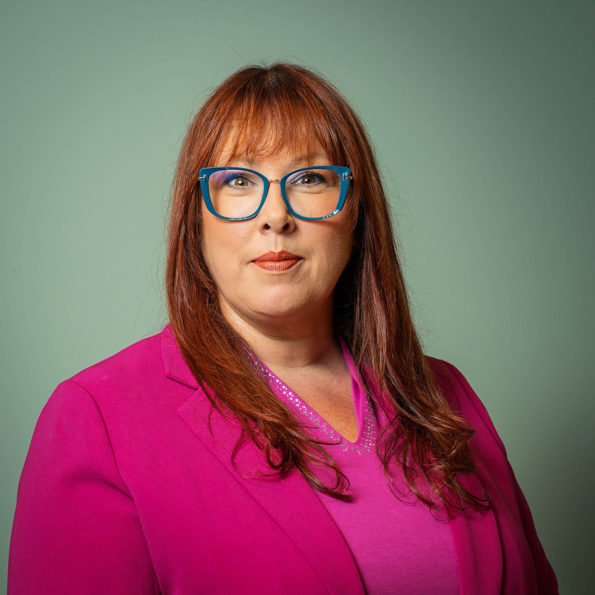 Een vrouw met rood haar, een blauwe bril en een roze blazer lacht naar de camera tegen een lichtgroene achtergrond.