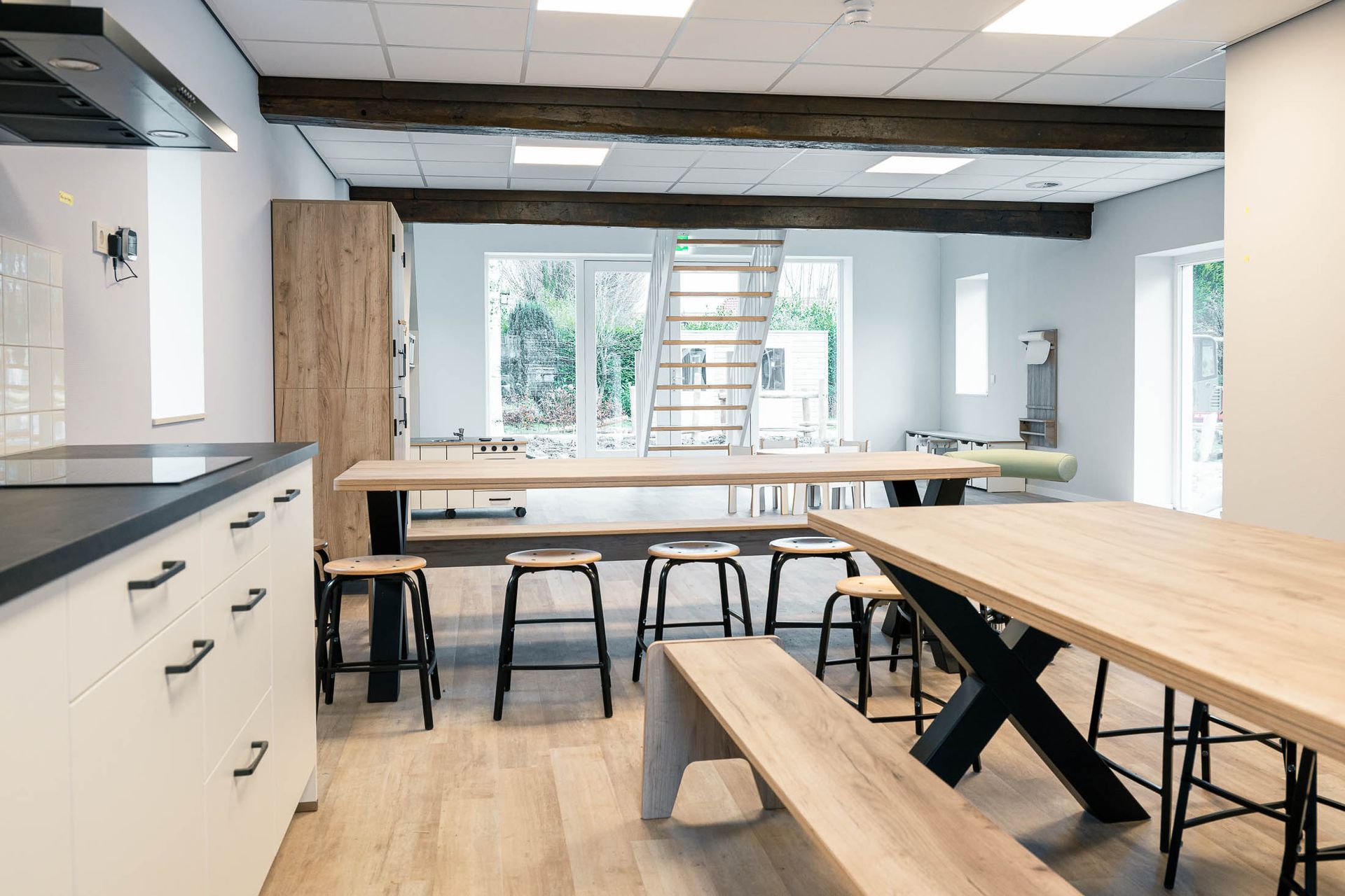 Keuken met lange houten tafels, zwarte krukken en witte kasten. Ramen geven toegang naar buiten.