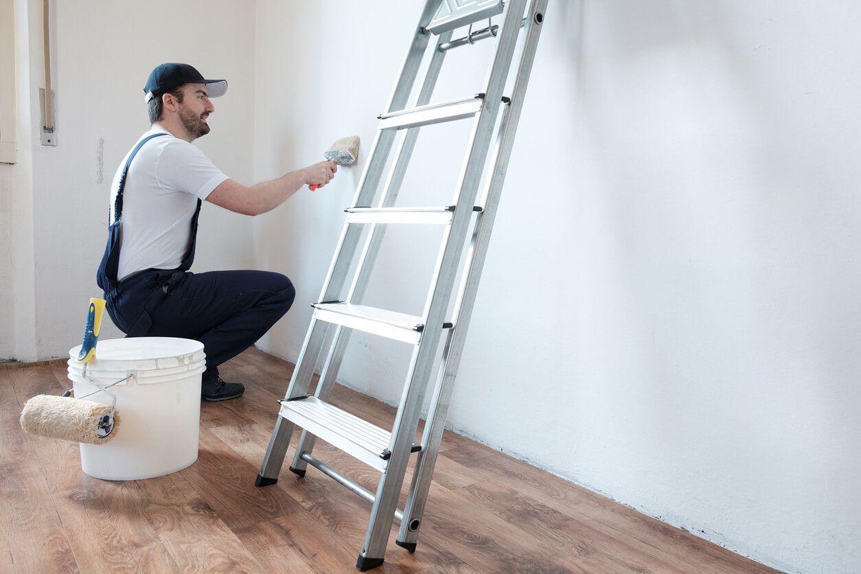 Painter using a roller to paint a white wall while kneeling beside a ladder and paint bucket.