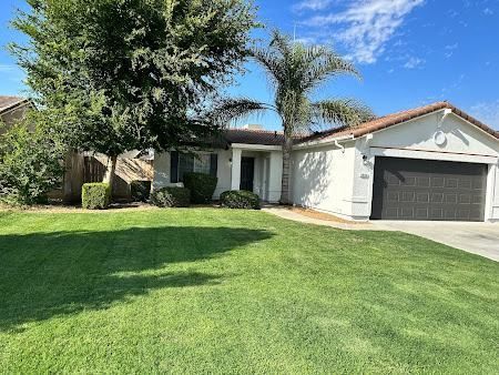 Single-story house with green lawn, trees, and a two-car garage. White exterior with a dark garage door.