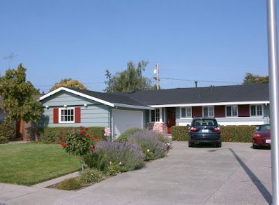 A light blue ranch house with red shutters, a driveway, and a dark blue car parked outside on a sunny day.