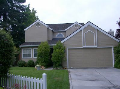 Beige two-story house with a white picket fence, green lawn, and a closed garage door.