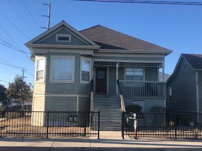 A two-story green house with a porch and stairs, set behind a black fence.