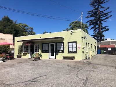 A light green building with a white door, black-framed windows, and a bench outside.