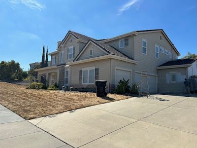Two-story beige house with brown roof, driveway, and dry grass on a sunny day.