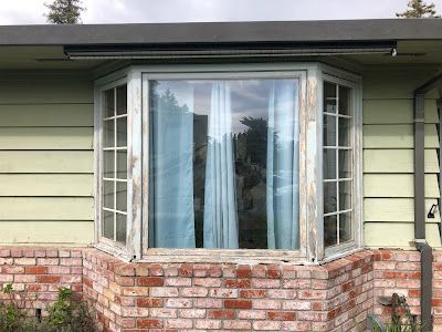 Bay window with peeling white paint on a brick and light green siding house. Blue curtains are visible.