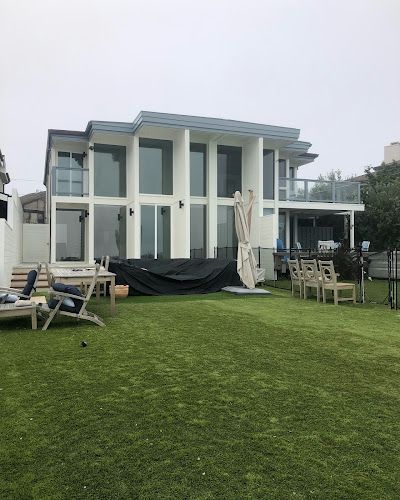 Modern white beach house with large windows, a green lawn, and outdoor seating. Overcast sky.