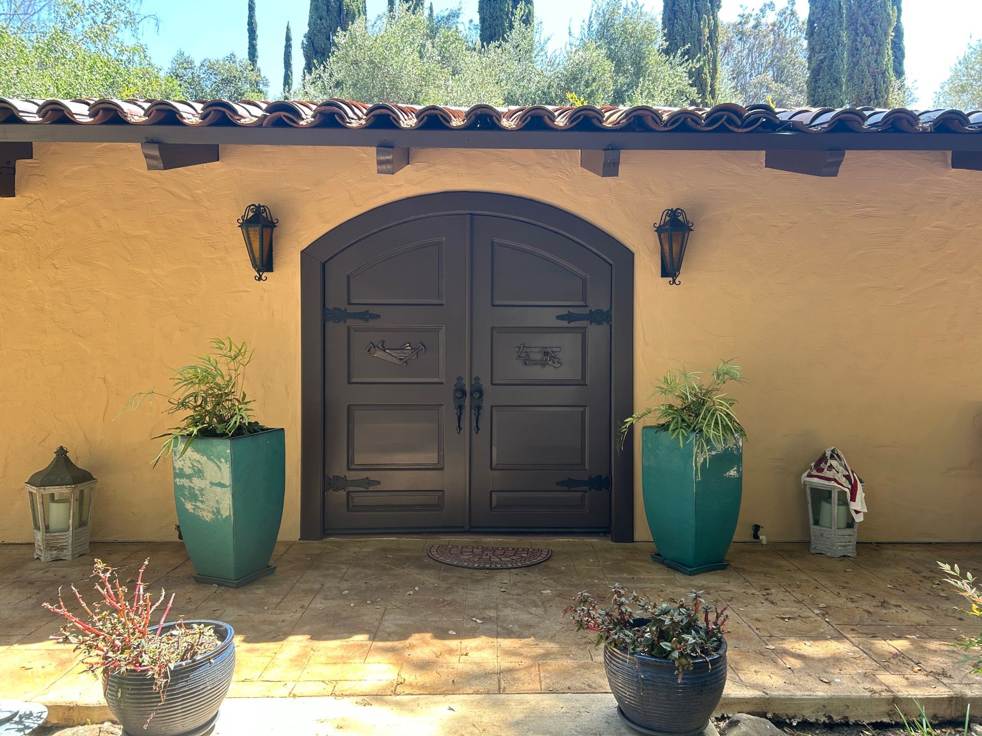 Double brown arched doors set in tan wall with terracotta roof, flanked by green planters.