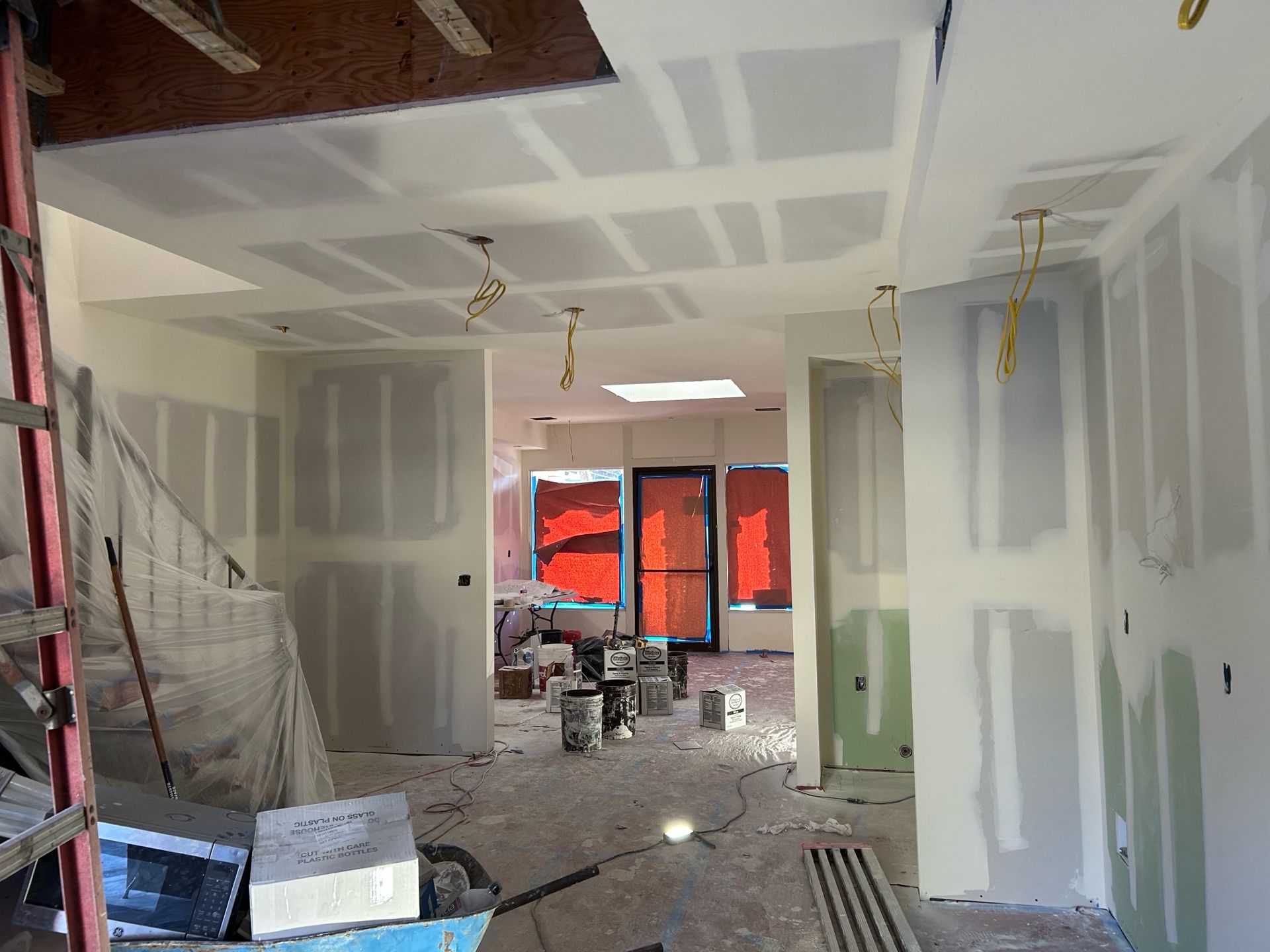 Interior of a building under construction, with drywall on walls and ceiling. Tools and supplies are scattered around.