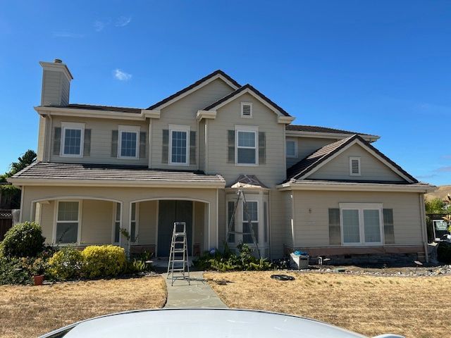 Two-story beige house with brown roof, shutters, and clear blue sky. Ladder near front door.