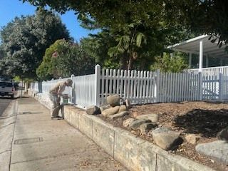 Person painting a white picket fence next to a concrete wall and sidewalk on a sunny day.