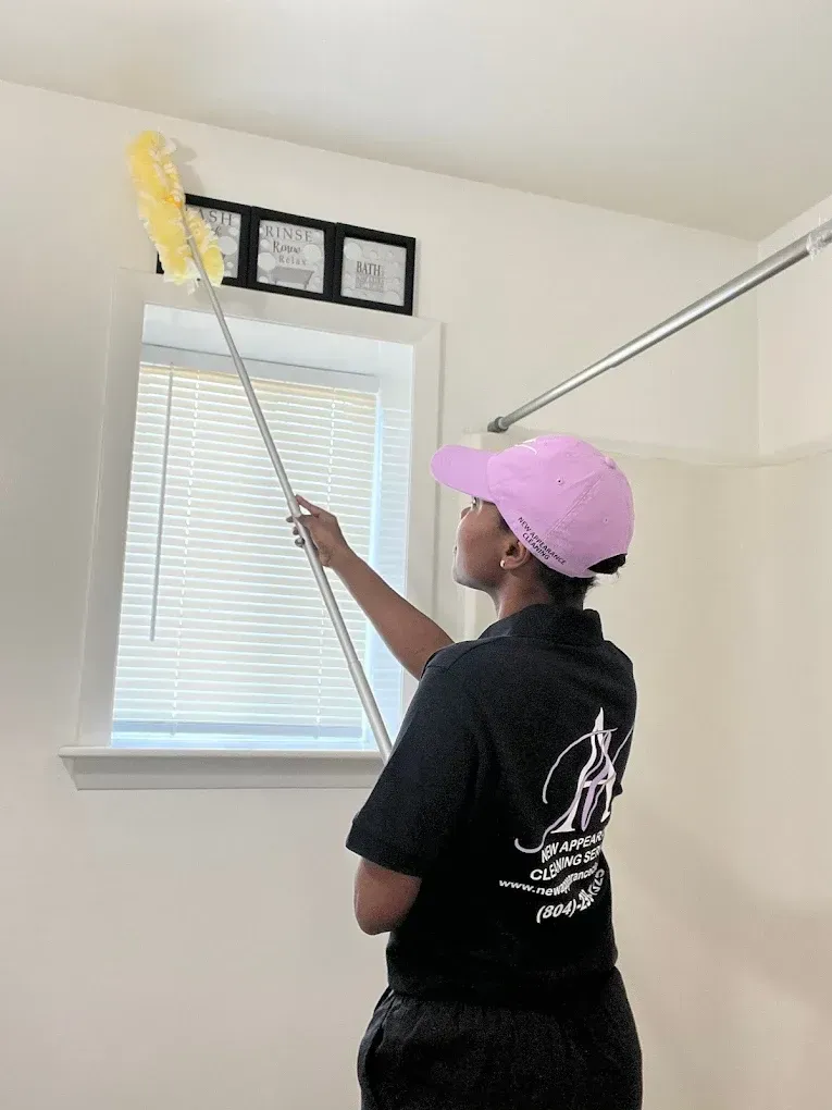 Person dusting above a window with a long-handled duster. They wear a pink cap and black shirt.
