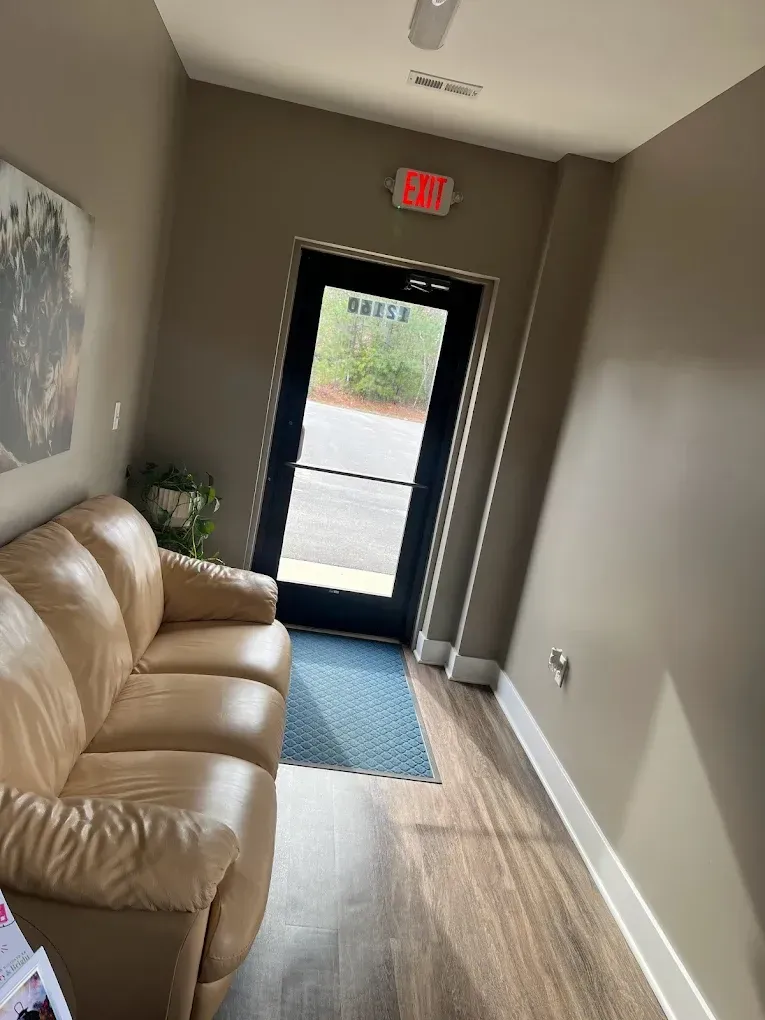A waiting area with tan couch, door, and exit sign. Light brown walls, wood-look floor, and a small rug.