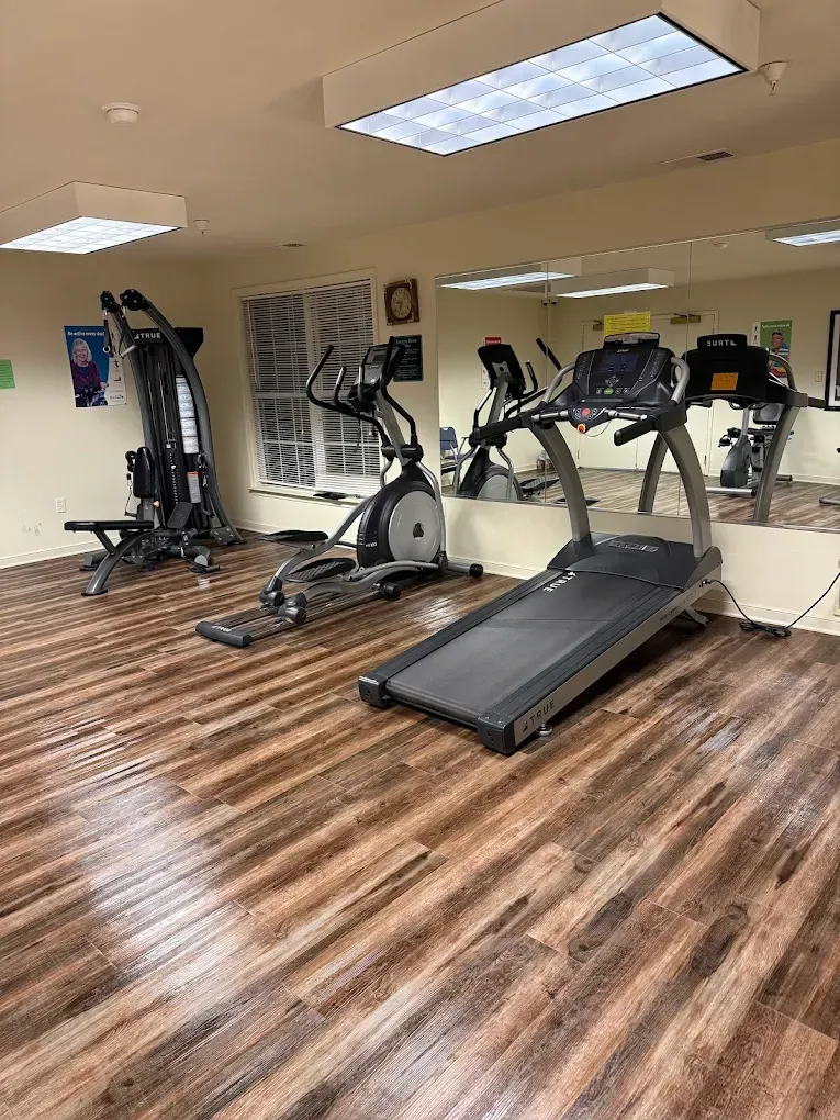 Gym with cardio machines on a wood-look floor, reflected in a large mirror.