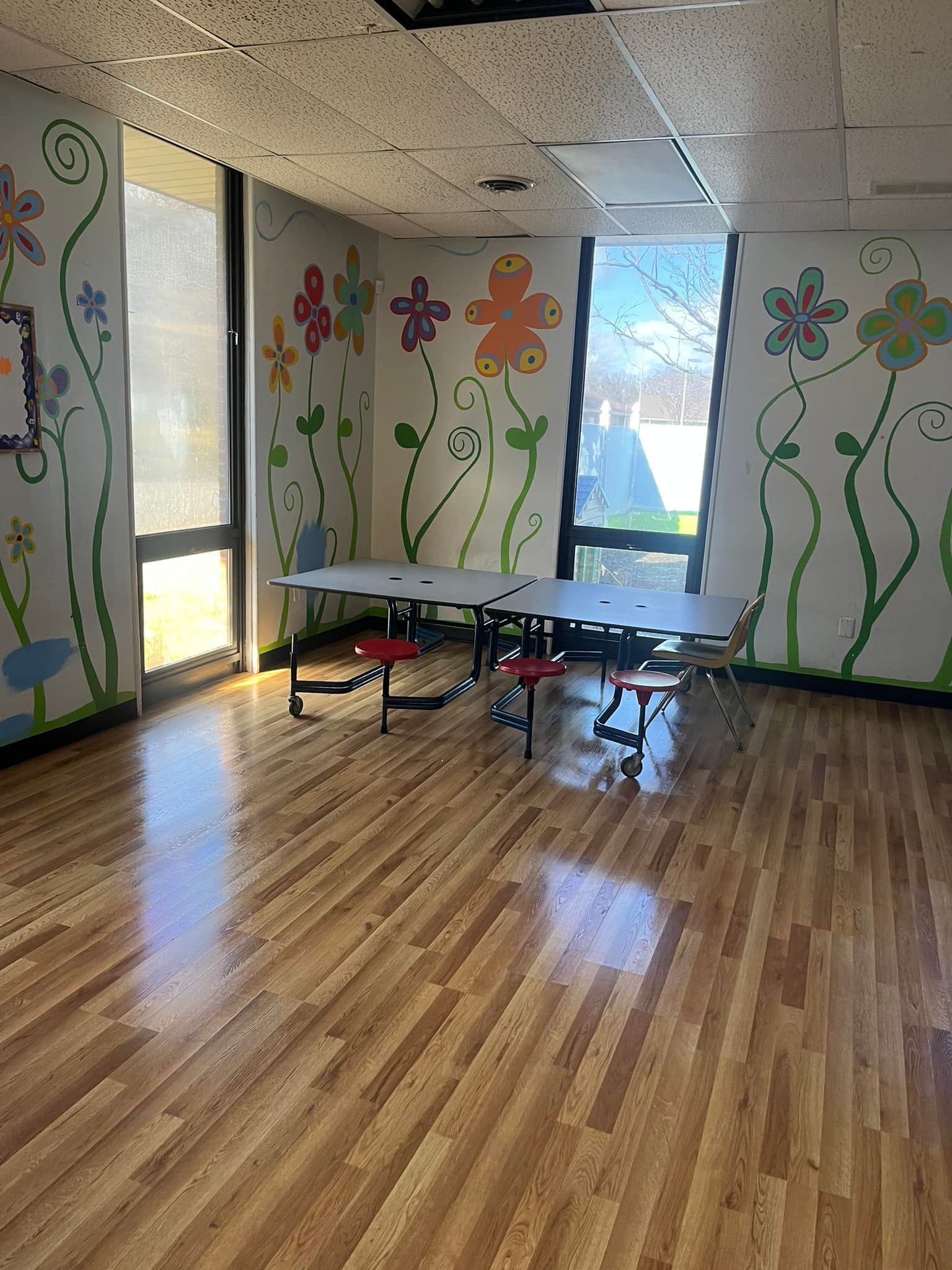 Room with floral wall art, tables, chairs, and large windows. Wooden floor, sunlight.