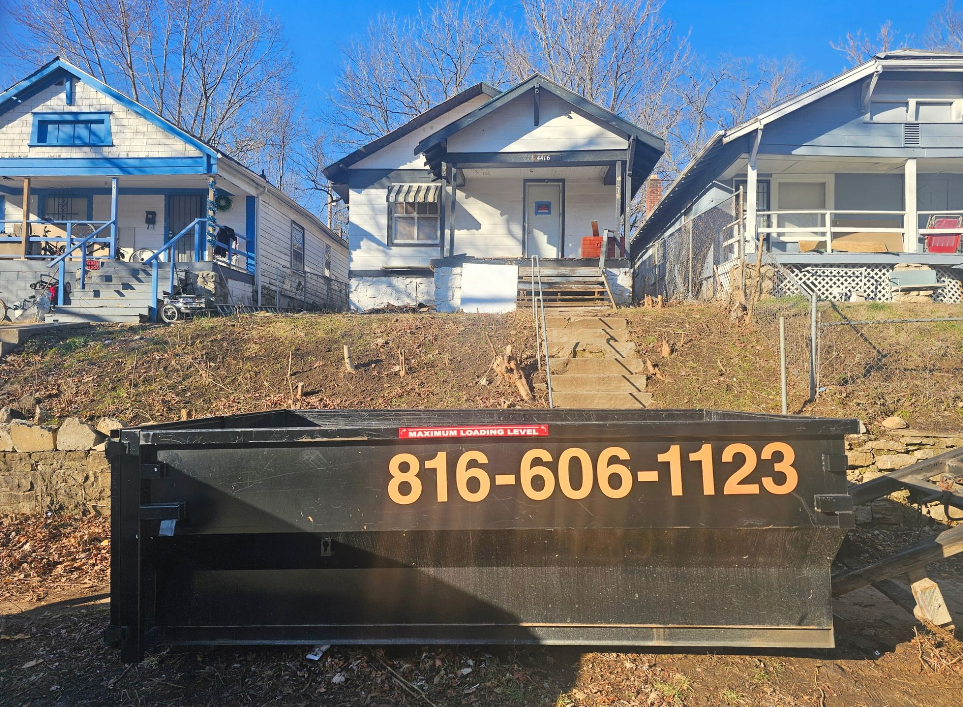 Yellow dumpster on a street next to a brick building and a gray truck.