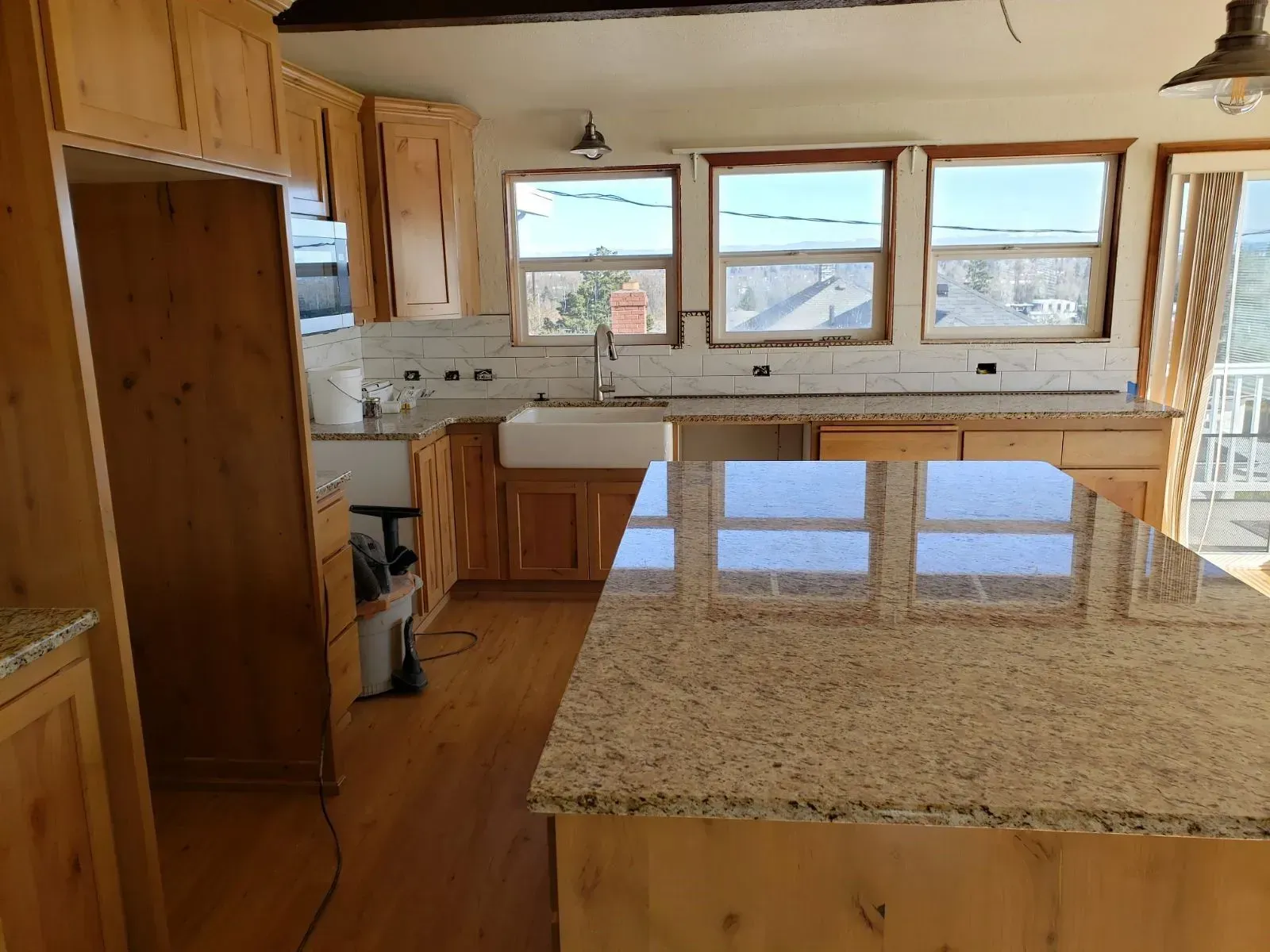 A kitchen with light wood cabinets, granite countertops, a farmhouse sink, and three windows overlooking the outdoors.