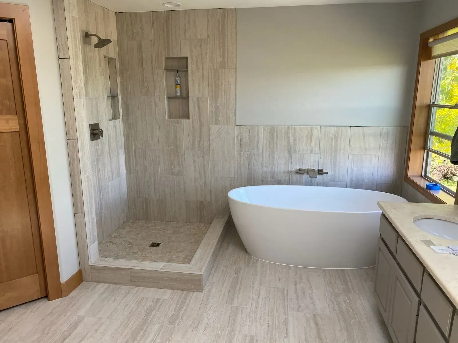 A modern bathroom with a walk-in shower featuring wood-look tile, a white freestanding tub, and light wood cabinetry.