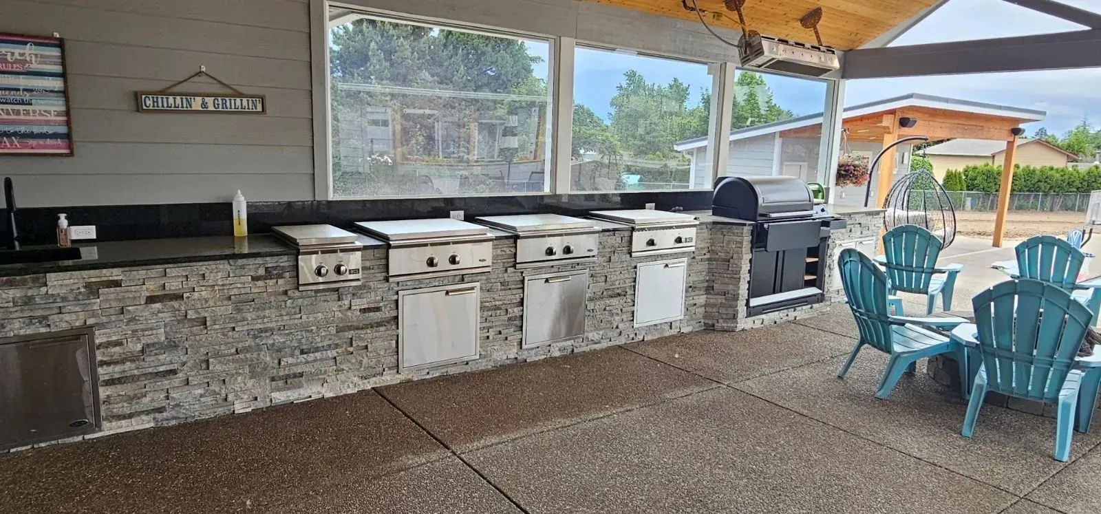 An outdoor kitchen with a stone-faced island, multiple grills, and turquoise Adirondack chairs on a concrete patio.