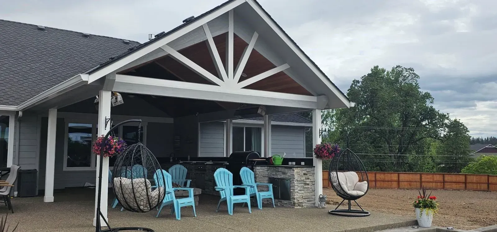 Covered patio with light blue chairs and two hanging basket chairs on a stone-paved yard under a cloudy sky.