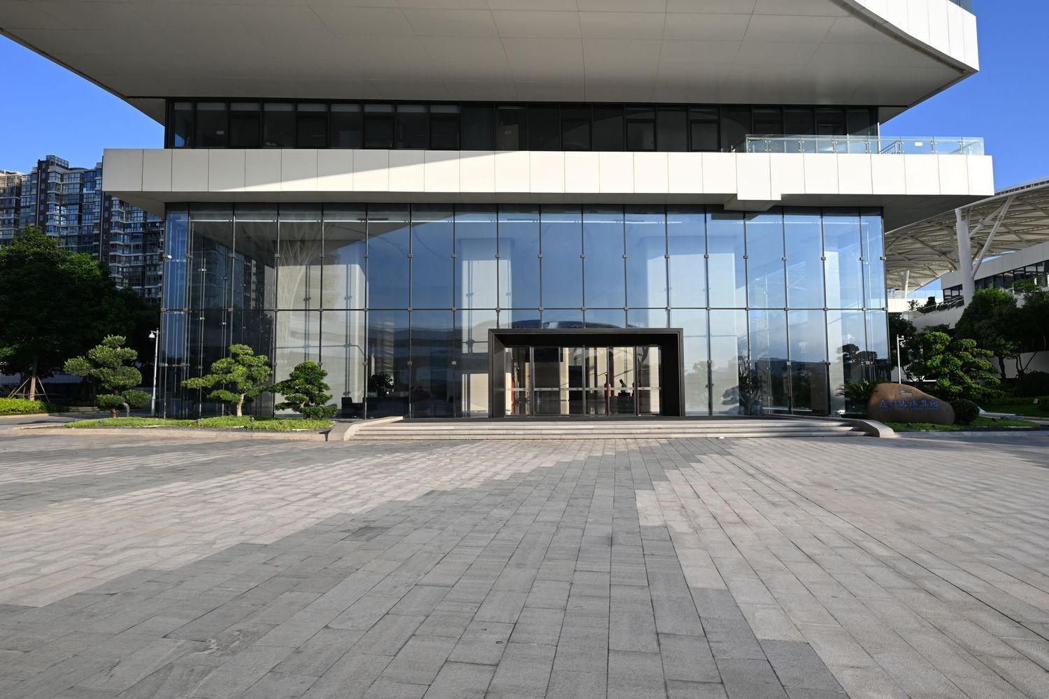 Modern building with a large glass facade, minimalist white overhang, and a wide paved plaza in front under a blue sky.