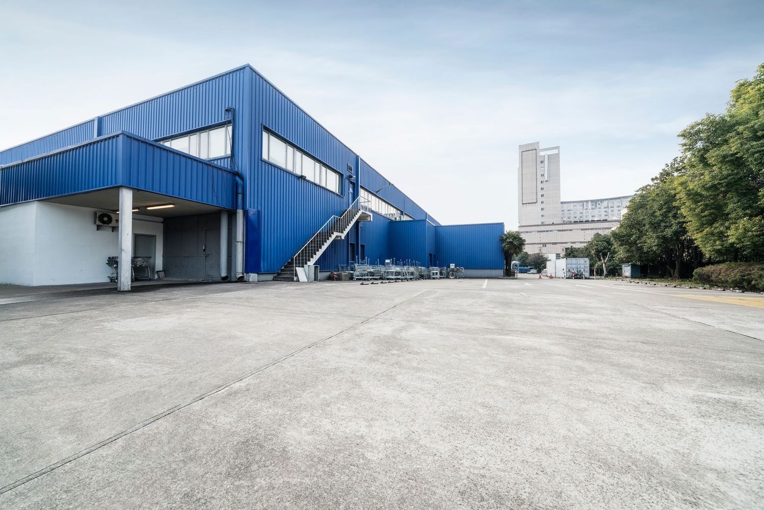 A large blue metal industrial warehouse with an exterior staircase and a spacious concrete lot under a clear sky.