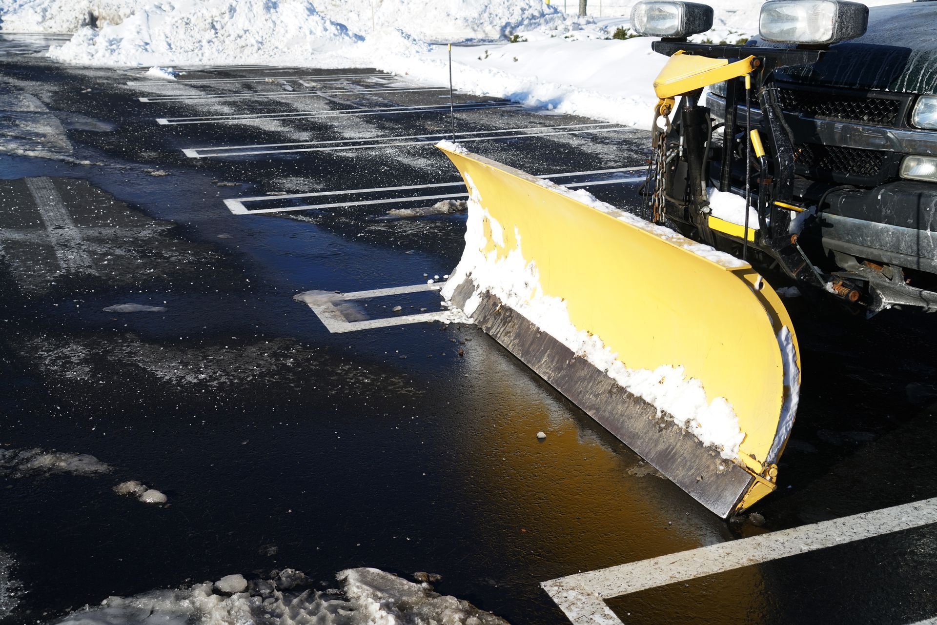 Yellow snowplow clearing snow from a black asphalt parking lot with white painted lines.