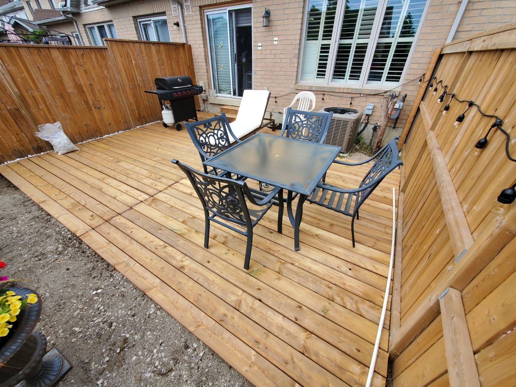 Wooden deck with a table and chairs, enclosed by a wooden fence, with a grill and string lights.