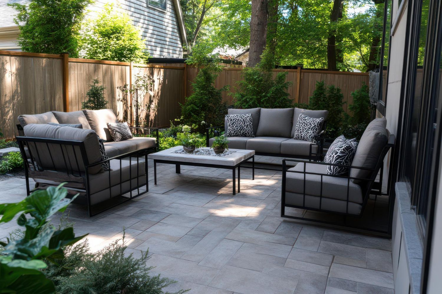 Patio with gray furniture, stone pavers, and wooden fence surrounded by greenery.