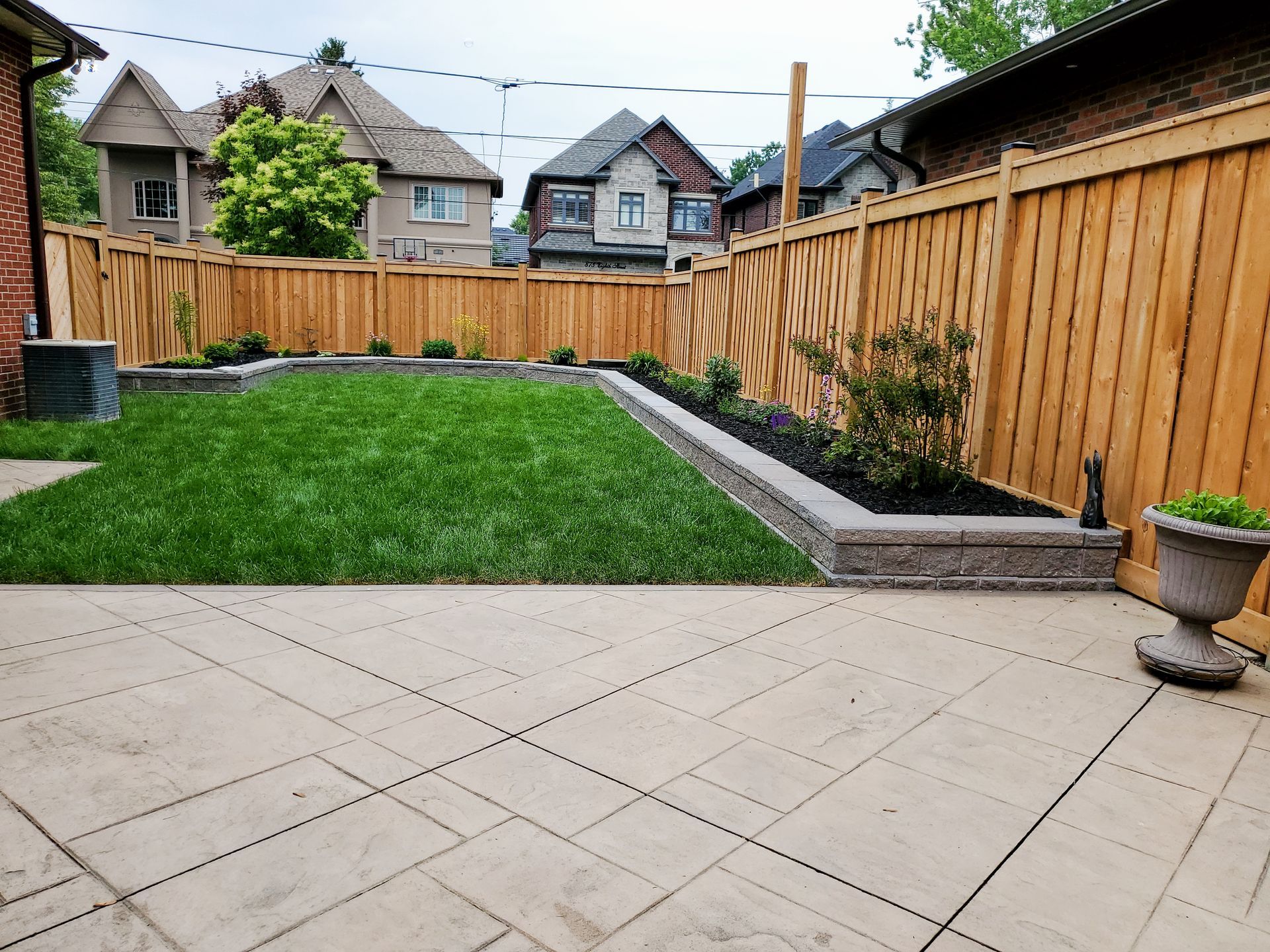 Backyard with grass lawn, paved patio, wooden fence, and neighboring houses beyond.