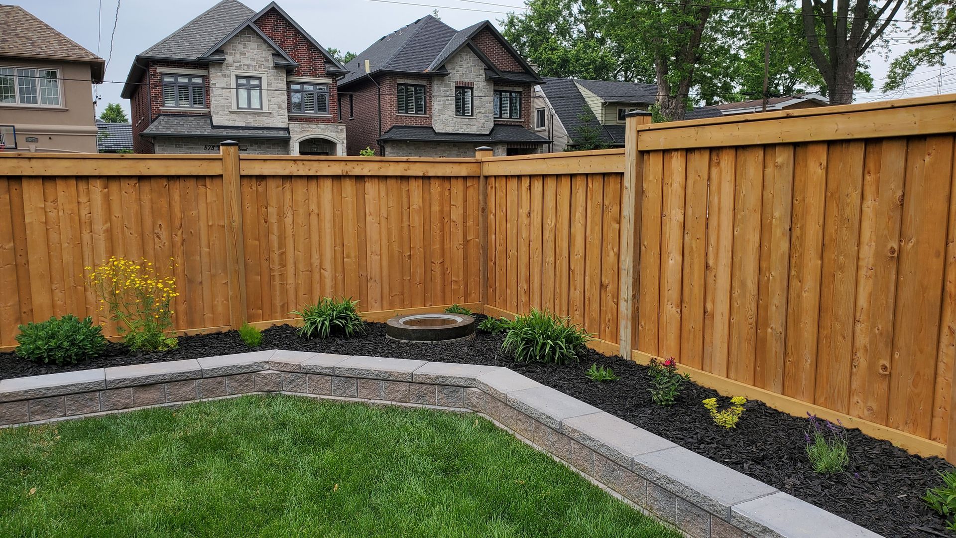 Backyard garden with curved stone edging, mulch beds, green grass, and a wooden privacy fence.