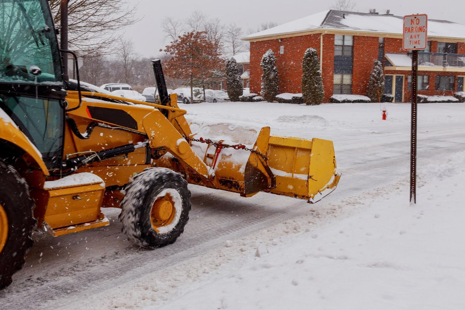 Yellow snowplow clearing snow from a residential street, buildings in the background.