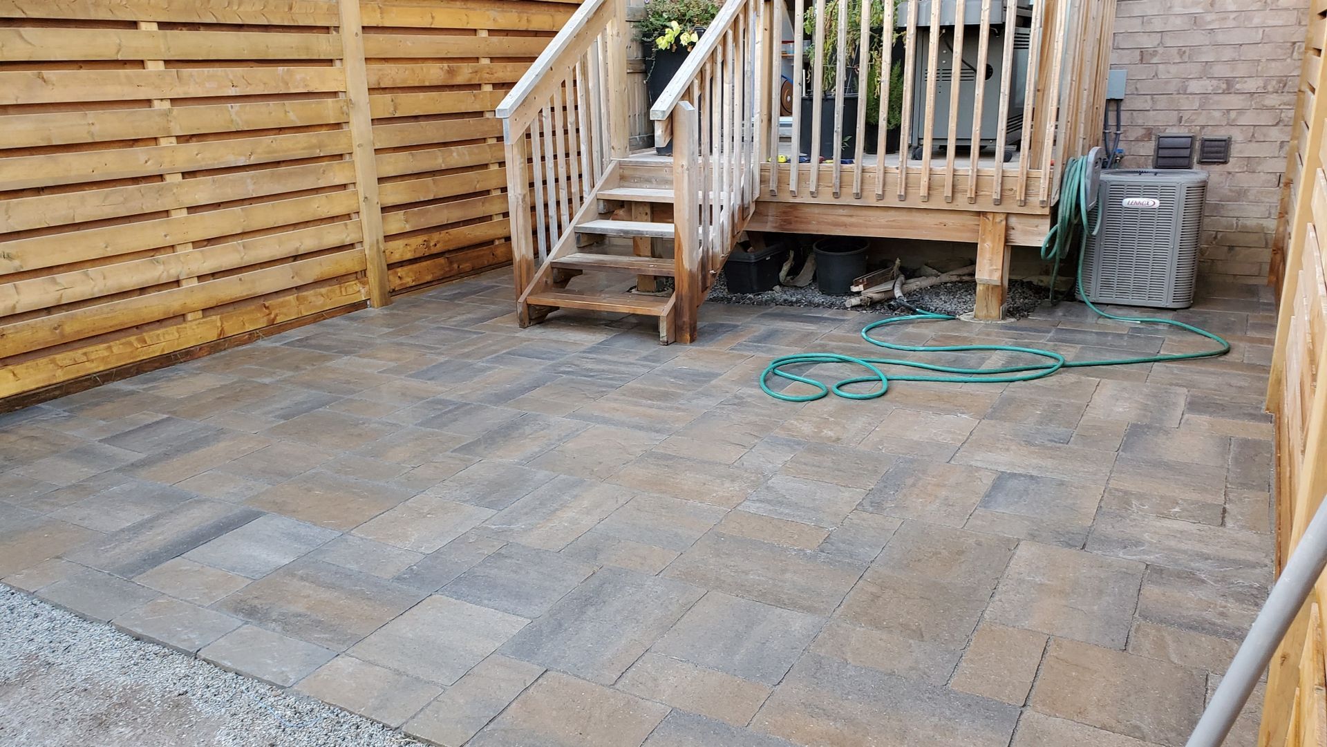 Paved backyard patio with wooden deck stairs, fence, and a coiled green hose