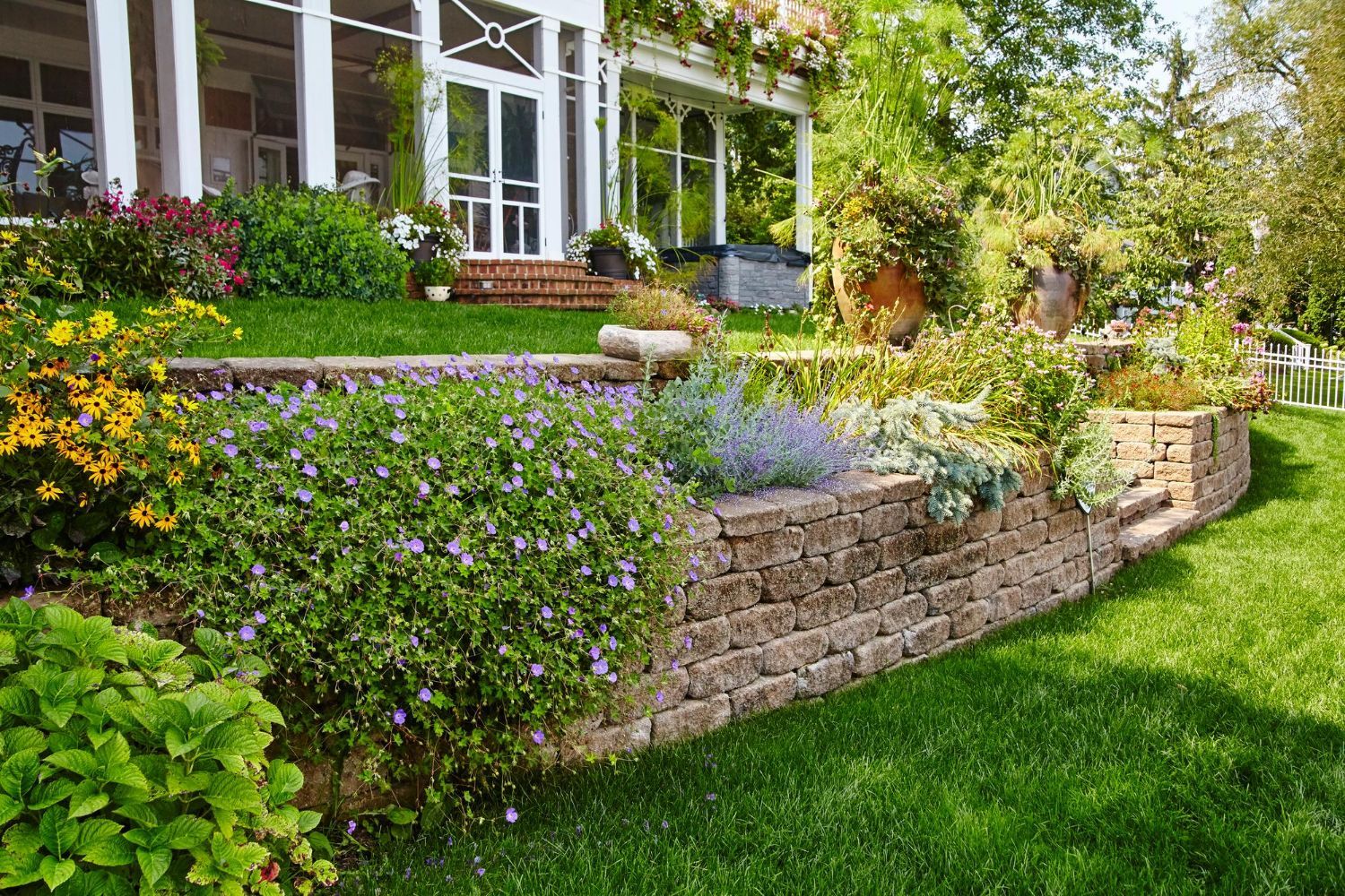 Stone retaining wall with flowers and lush greenery in a sunny garden. House in the background.