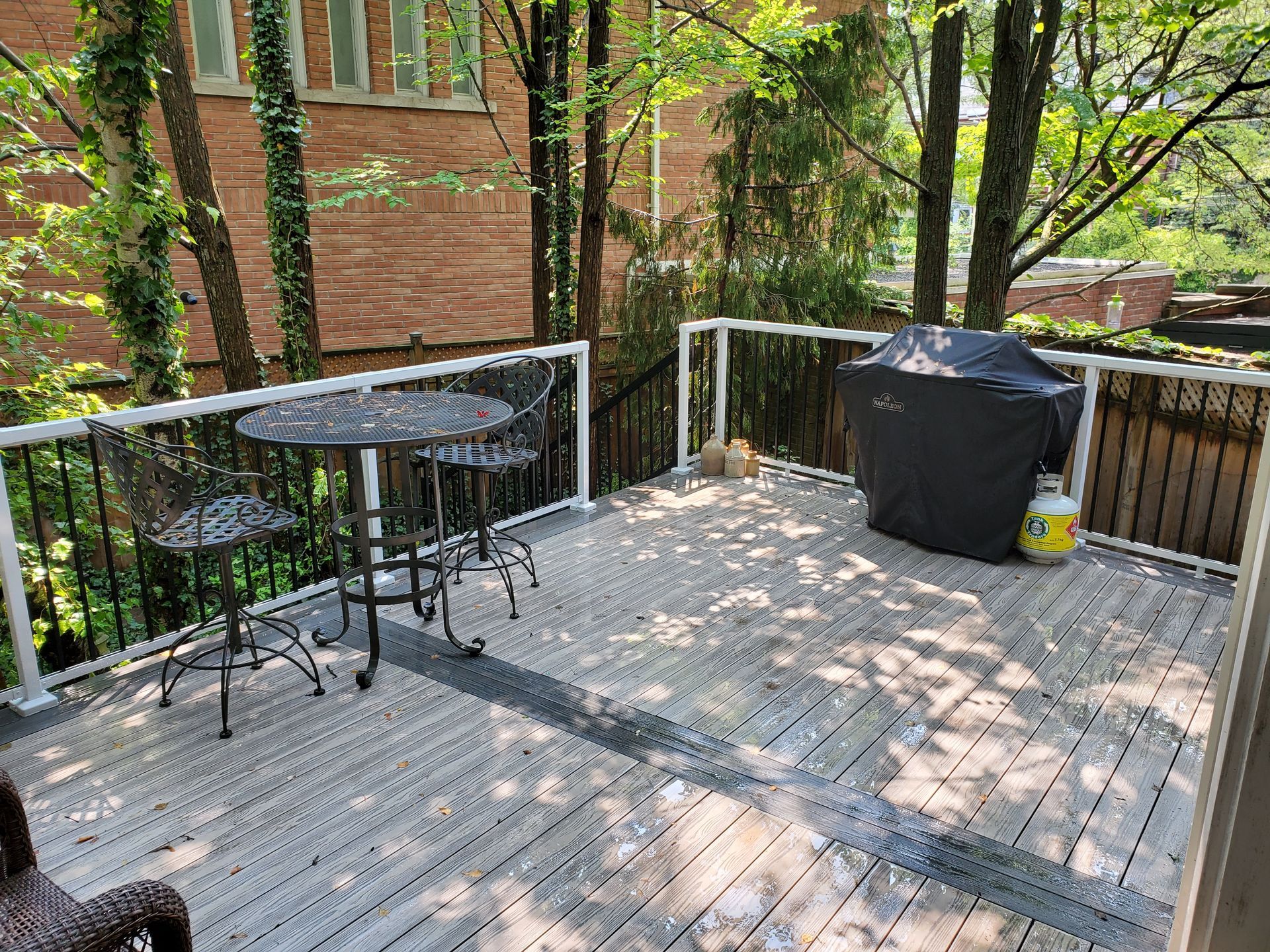 Deck with table, chairs, covered grill, and propane tank. Surrounded by trees and a building.