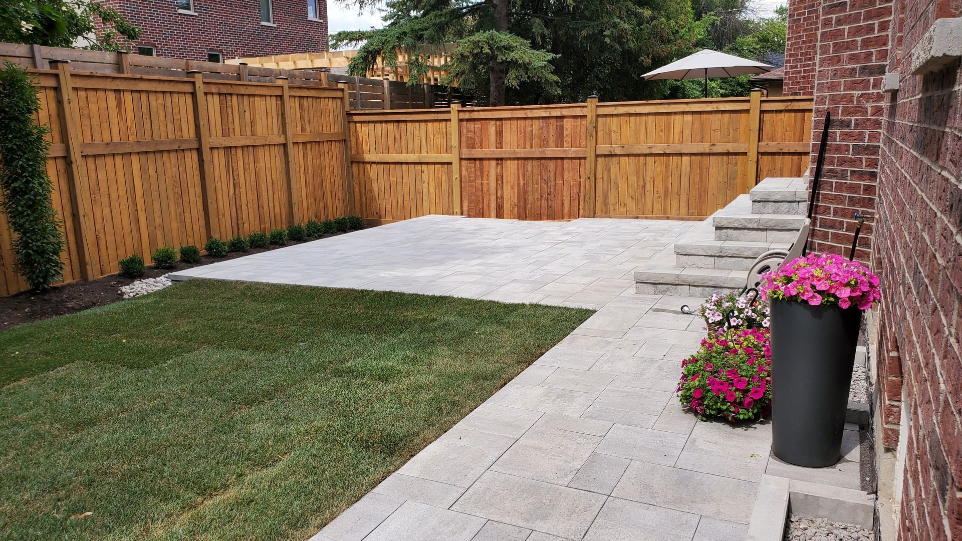 Backyard patio with light stone pavers, green lawn, wooden fence, and potted pink flowers beside a brick wall