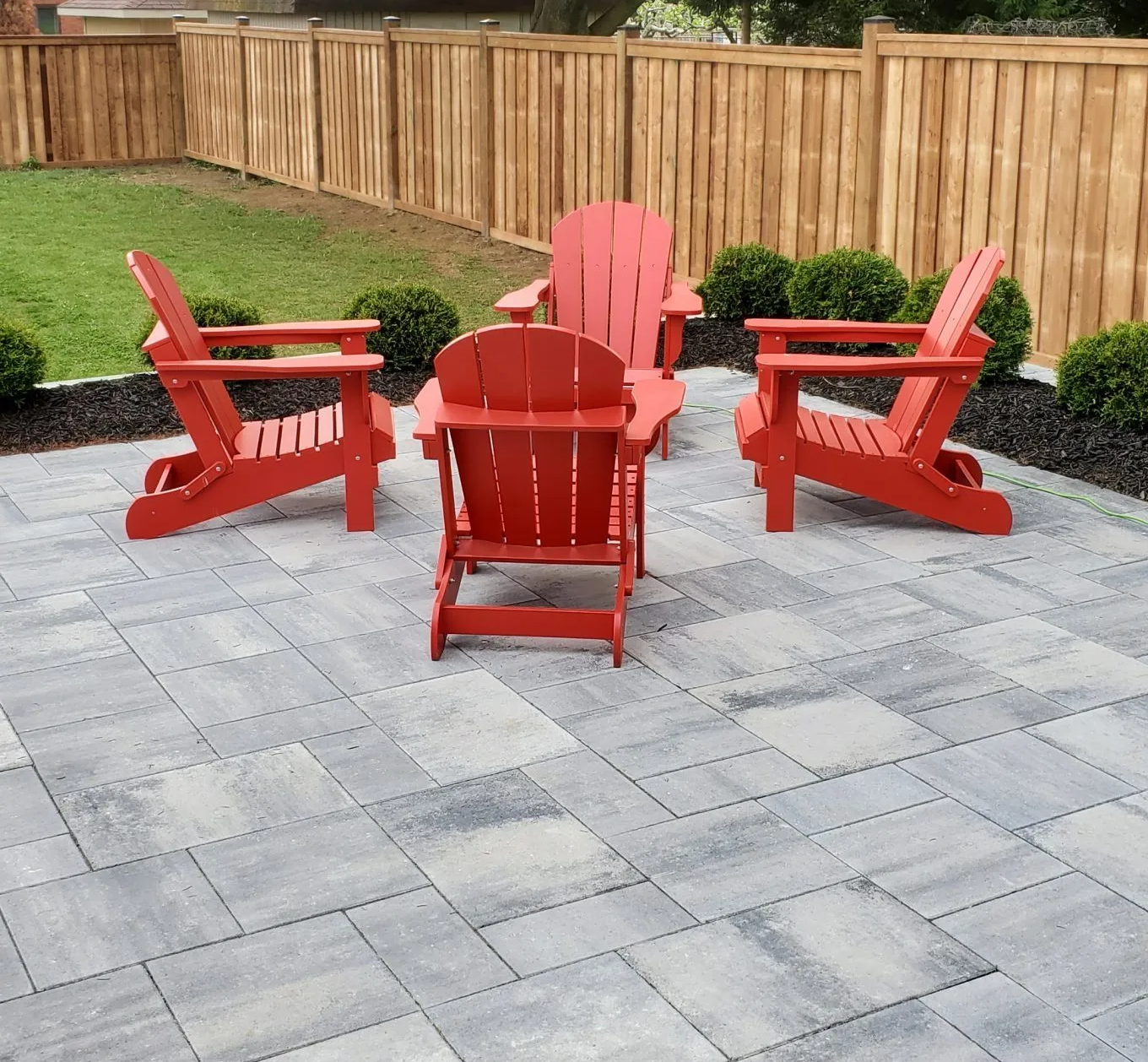 Four red Adirondack chairs on a gray paver patio, surrounded by greenery and a wooden fence.