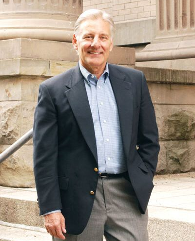 Man in navy blazer and grey pants smiles, standing near building steps.