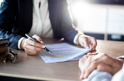 Person in blazer pointing at document on table; another person’s hands visible.