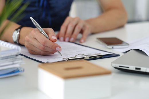 Woman writing on clipboard at a desk; watch, phone, laptop, and box are nearby.
