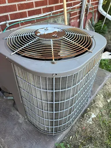 Air conditioning unit with rusty top grille, next to brick wall and grass.