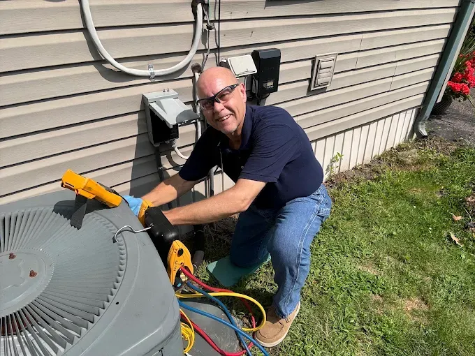 Man fixing an AC unit. He's wearing blue and jeans and smiling. Outside near siding and grass.